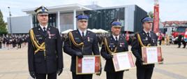 Czterech funkcjonariuszy Państwowej Straży Pożarnej w mundurach galowych pozuje do zdjęcia, trzymając w rękach dyplomy wyróżnień. W tle widać strażaków w szyku oraz nowoczesny budynek z przeszkloną fasadą.