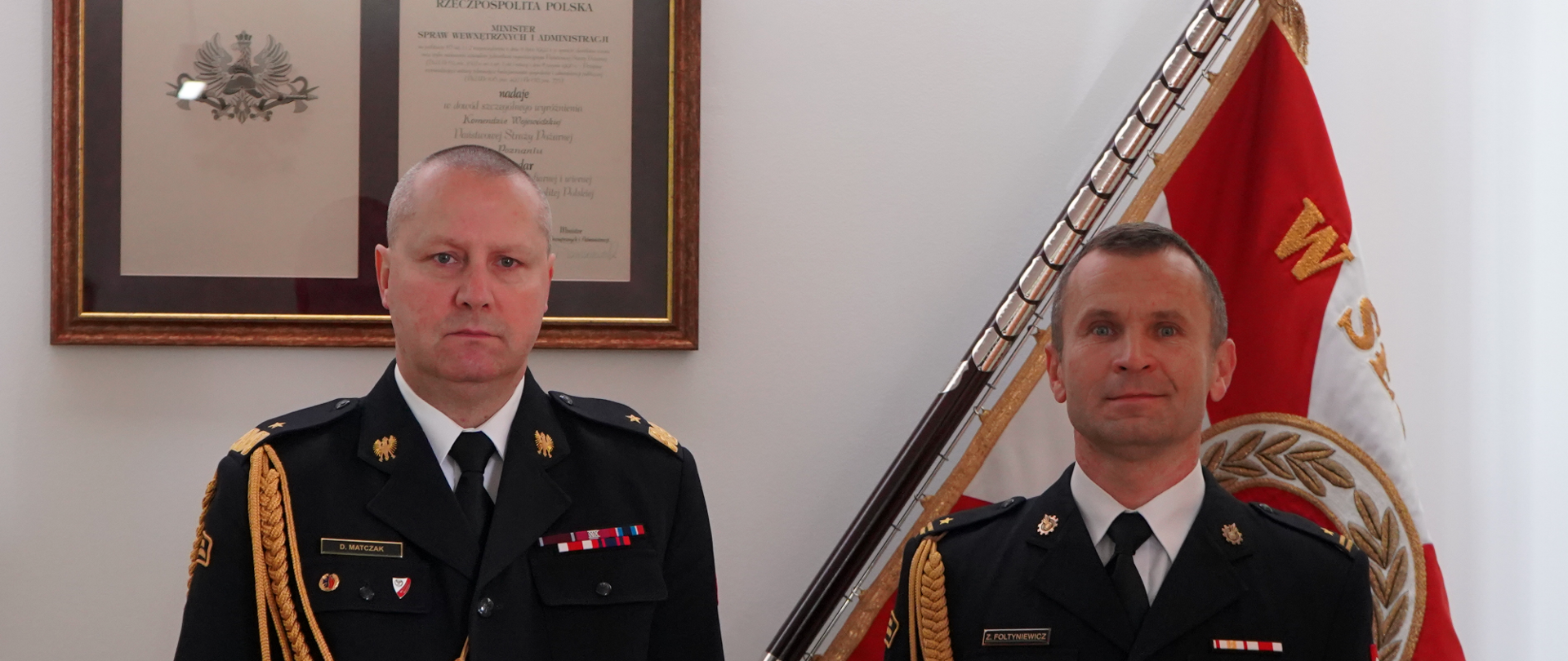 dwóch strażaków w mundurach ze sznurem stoi obok siebie, w tle sztandar