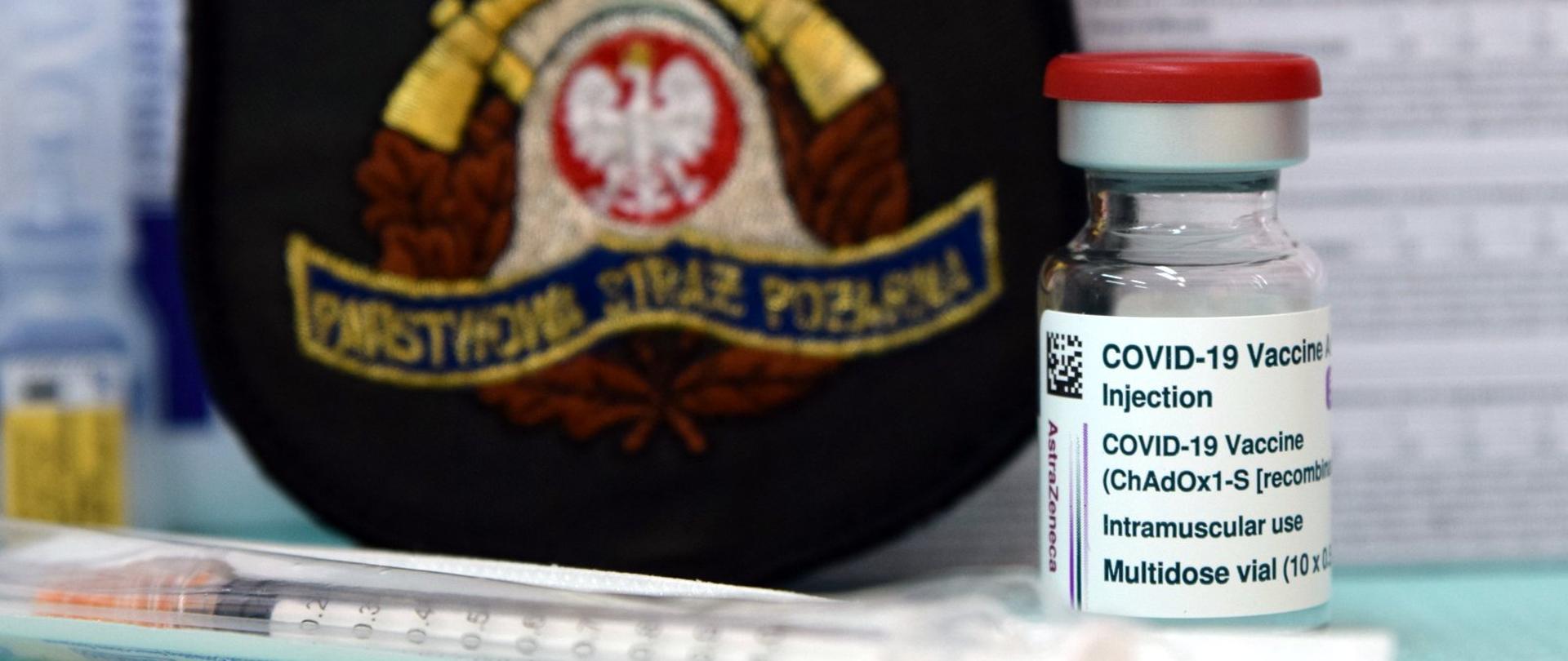 Na zdjęciu widoczna jest fiolka ze szczepionką przeciw COVID-19 przed nią leży strzykawka jednorazowa. W tle emblemat Państwowej Straży Pożarnej