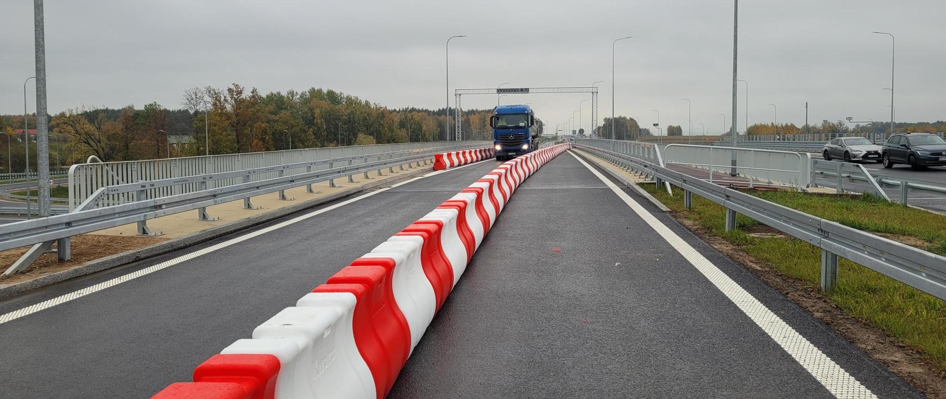 Nowo wybudowany odcinek drogi lub mostu z ciemną nawierzchnią asfaltową. Przez środek jezdni biegnie długi ciąg czerwono-białych barier separacyjnych. W oddali widoczna jest niebieska ciężarówka jadąca w kierunku aparatu. Po obu stronach drogi znajdują się metalowe bariery ochronne i wysokie latarnie. W tle widoczne są drzewa o jesiennych barwach pod pochmurnym, szarym niebem.