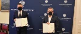 Zdjęcie przedstawia dwóch elegancko ubranych mężczyzn, trzymających w rękach umowy. Pozują oni na tle granatowej ścianki wizerunkowej Wojewody Mazowieckiego, opatrzonej kilkoma logotypami Wojewody Mazowieckiego (stanowiącymi Godło Polski oraz napis "Wojewoda Mazowiecki").