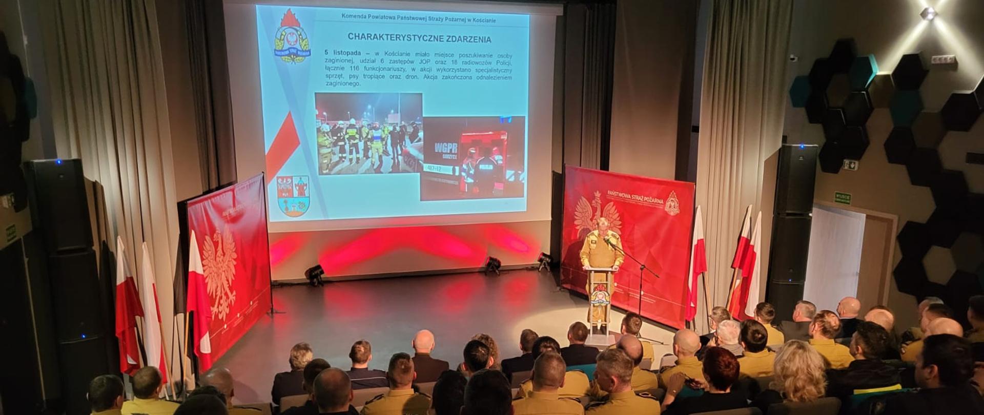 Kościańscy strażacy podsumowali 2025 rok. Strażacy i zaproszeni goście zebrani na sali podczas odprawy służbowej. Funkcjonariusz przemawia, w tle ekran z prezentowanym materiałem. 