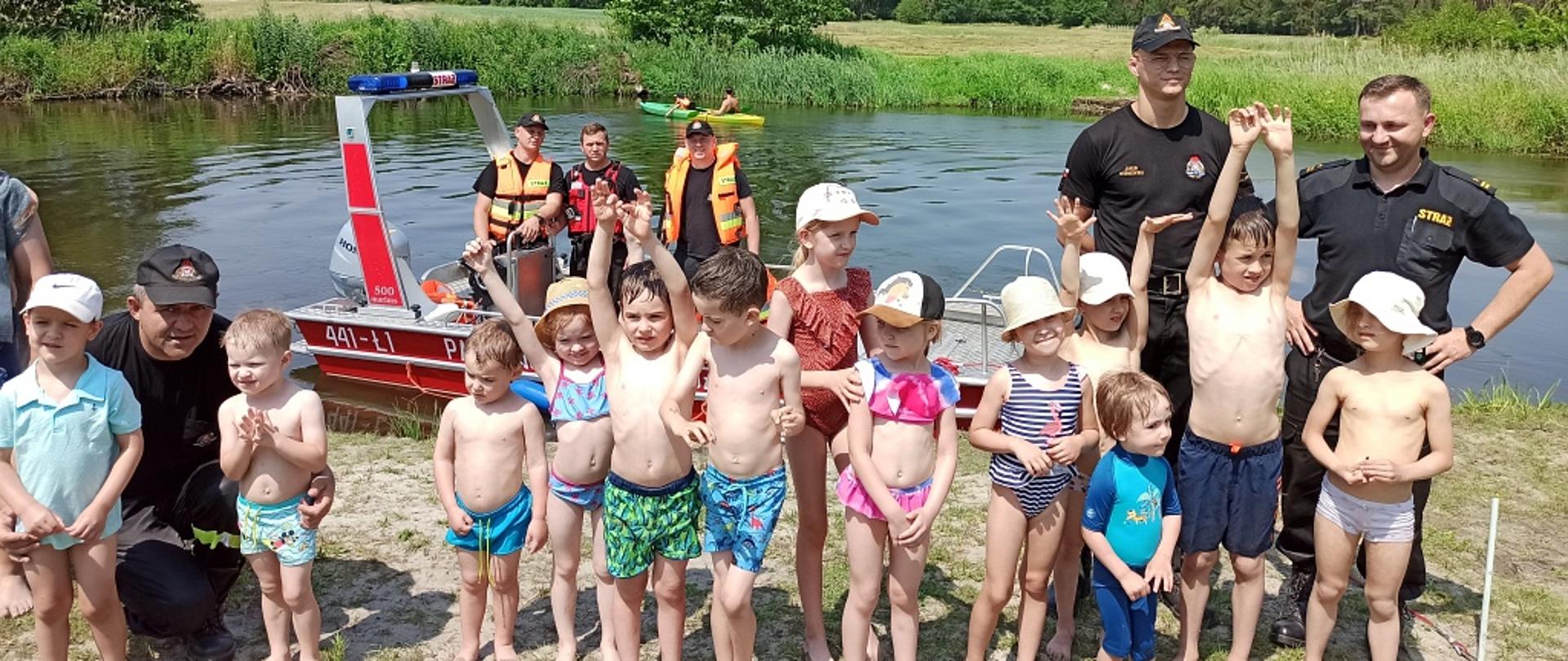 Na zdjęciu widać grupę dzieci wraz ze strażakami, w tle strażacy na łodzi na rzece