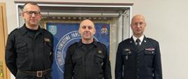 Trzech mężczyzn w mundurach Państwowej Straży Pożarnej stoi przodem ustawieni do wspólnego zdjęcia, za nimi widoczna jest gablota w której wisi sztandar Komendy powiatowej PSP w Pleszewie