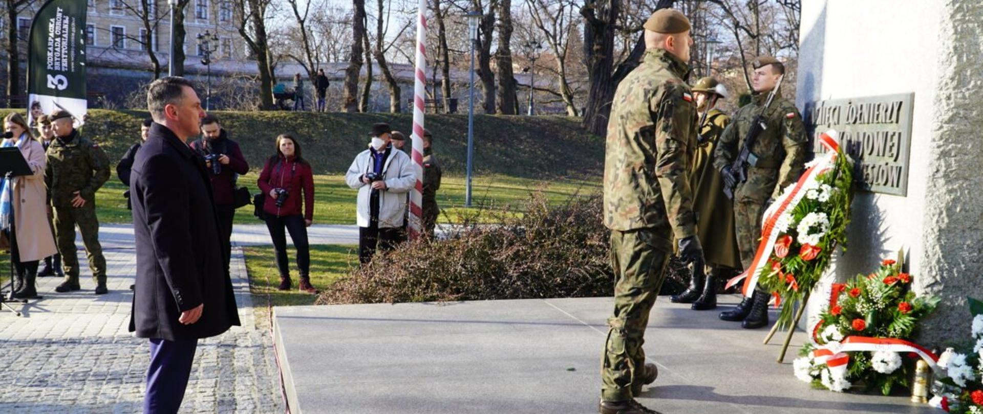 Dyrektor generalny Podkarpackiego Urzędu Wojewódzkiego w Rzeszowie składa kwiaty pod pomnikiem poświęconym pamięci żołnierzy Armii Krajowej w Rzeszowie. Przed nim żołnierz, w tle fotoreporterzy i uczestnicy uroczystości.