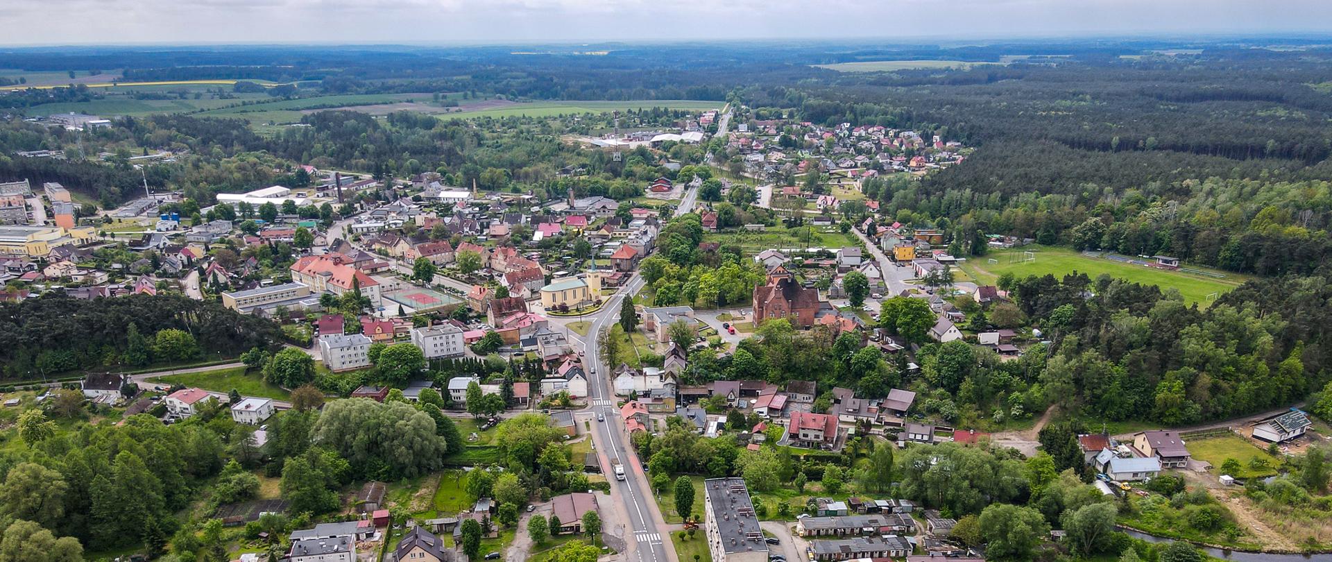 Na zdjęciu widać niewielkie, zielone miasteczko sfotografowane z lotu ptaka, otoczone lasami i polami. Przez jego środek biegnie główna droga, a zabudowa jednorodzinna i centrum miejscowości harmonijnie wtapiają się w otaczającą zieleń.