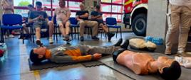 W strażnicy pożarnej trwa szkolenie z zakresu pierwszej pomocy. Na podłodze leżą fantomy osoby dorosłej i dziecka wykorzystywane do ćwiczeń ratowniczych. W tle uczestnicy siedzą na krzesłach i obserwują zajęcia, a obok widoczny jest wóz strażacki. Zdjęcie przedstawia praktyczną część szkolenia dotyczącego ratowania życia.
