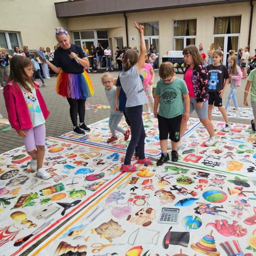 Grupa dzieci stoi na planszach posiadających kolorowe symbole różnych przedmiotów.