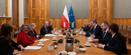 Duża sala ze ścianami w zielony wzorek, przy drewnianym stole siedzi kilkanaście elegancko ubranych osób. Po prawej stronie delegacka Ukrainy po lewej kierownictwo MEN. Na stole rozłożone notatki oraz ustawione napoje. W tle flagi Polski i UE