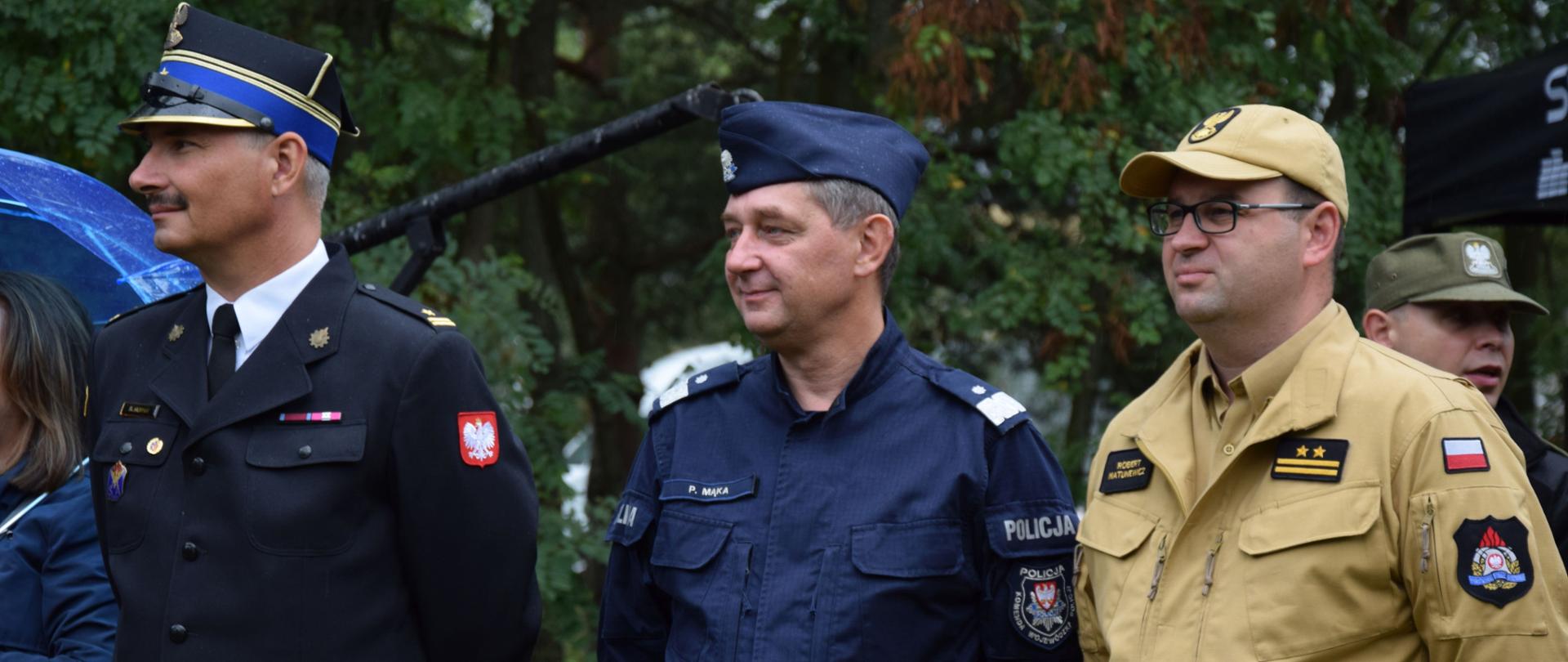 trzech mężczyzn stoi w szeregu, z prawej strażak w jasnym mundurze i w czapeczce oraz w okularach, w środki policjant w niebieskim mundurze i furażerce, po lewej strażak w mudnurze wyjściowym i rogatywce, w tle drzewa