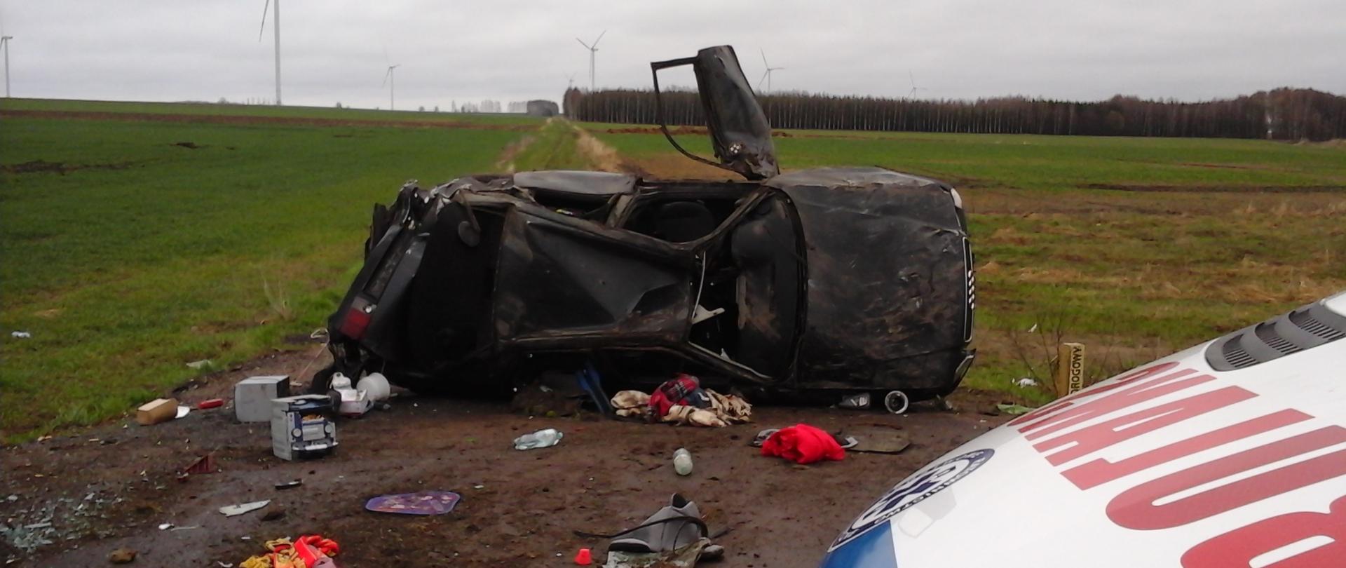 Ciemny samochód osobowy marki Audi leżący na boku na poboczu drogi. Nadwozie samochodu jest zniszczone, okna powybijane. W tle łąki i las.