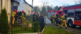 Zdjęcie przedstawia wejście do budynku, wewnątrz którego wybuchł pożar.
Na zewnątrz znajdują się ratownicy oraz samochody gaśnicze. 