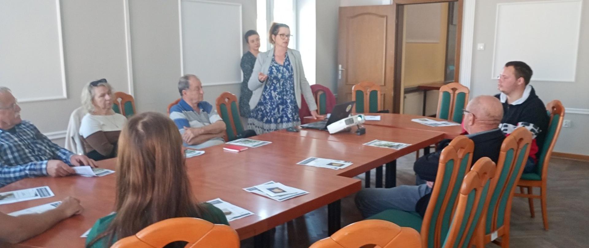 Przy stole siedzą uczestnicy spotkania i słuchają wykładu prezentowanego przez stojącą panią inspektor 