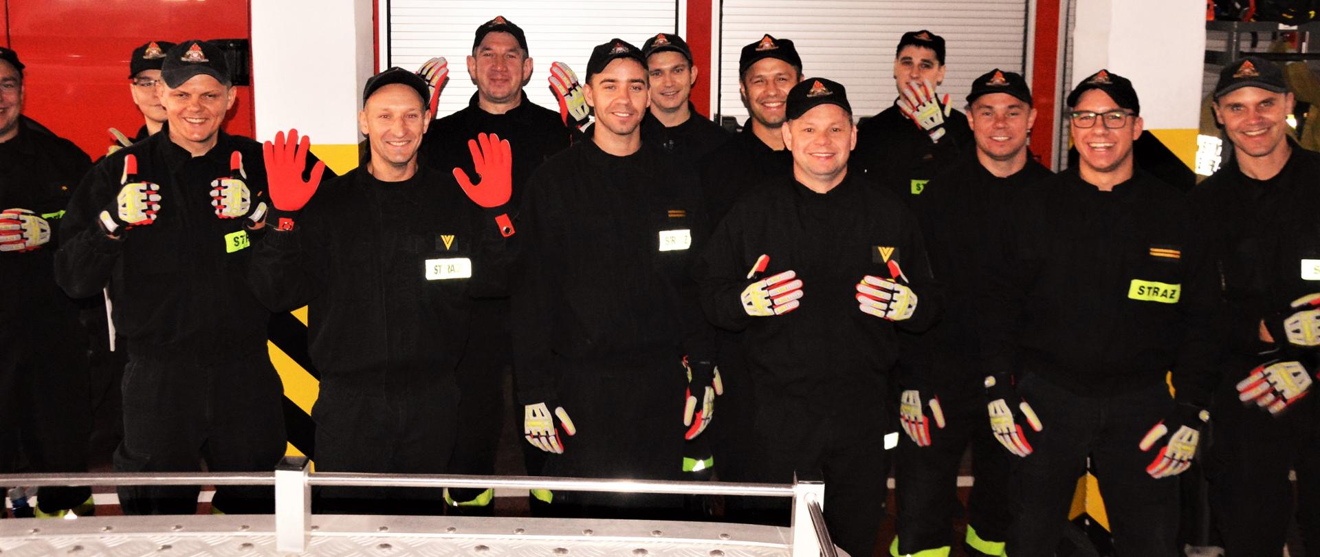 Prezent dla strażaków. Garaż Komendy Powiatowej PSP w Rawiczu. Na tle wozu strażackiego stoją strażacy z dwóch zmian służbowych. Każdy z nich prezentuje nowe rękawice do ratownictwa technicznego. 