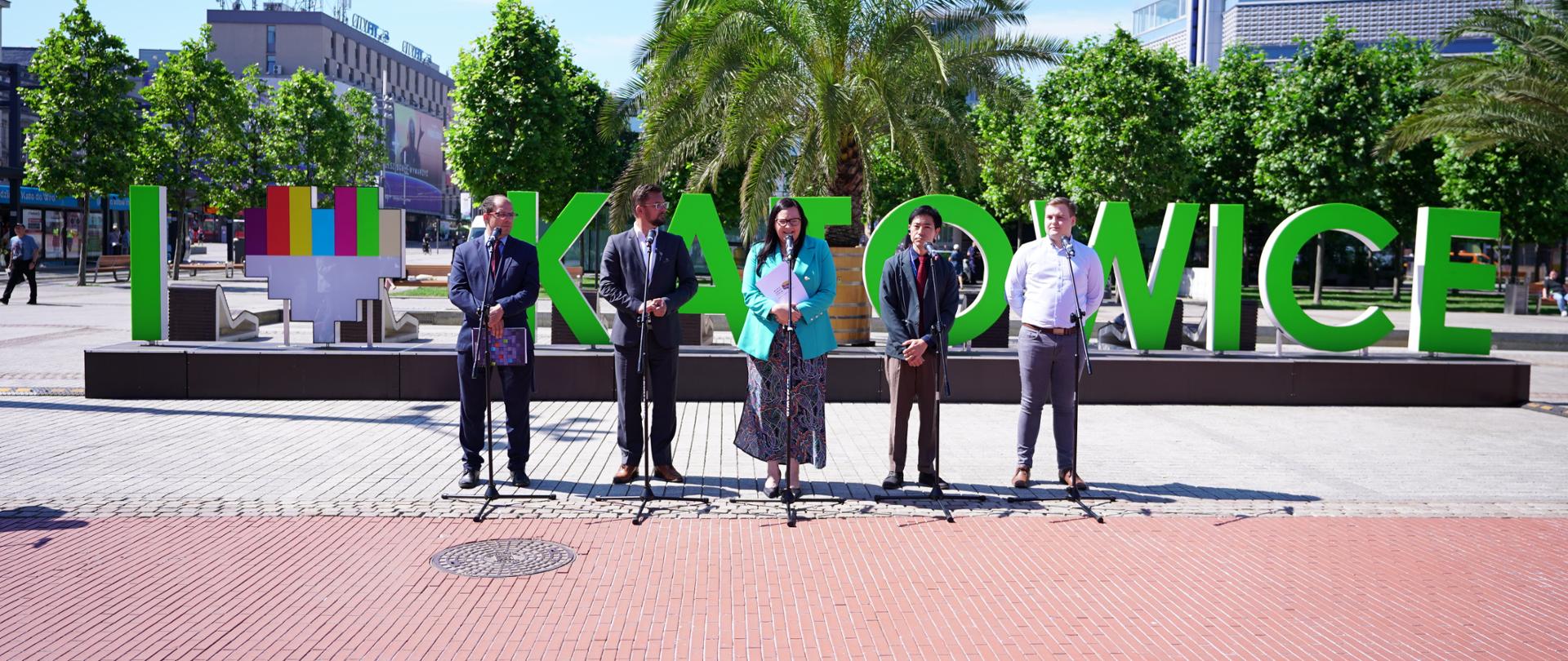 Na zdjęciu przemawiający na konferencji prasowej stojący na rynku w Katowicach. Za uczestnikami duży zielony napis "Katowice", przed uczestnikami stojące mikrofony. W tle drzewa.
