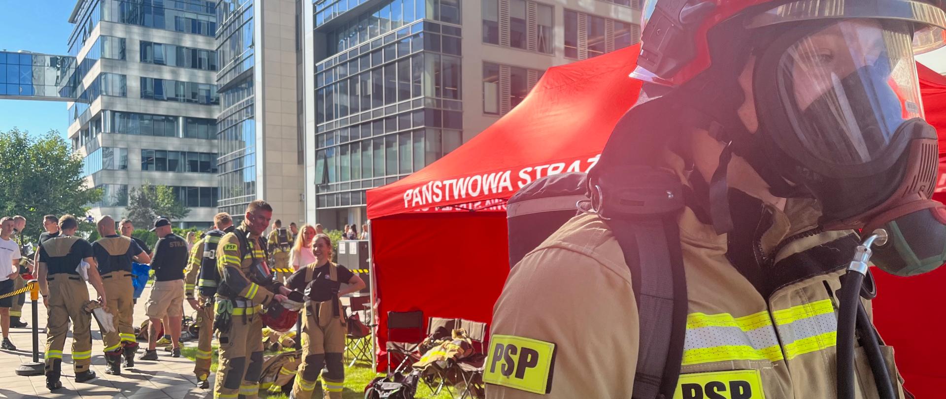 Strażak PSP w aparacie ochrony dróg oddechowych (AODO) stoi na pierwszym planie przed czerwonym namiotem z napisem „Państwowa Straż Pożarna”, w tle widoczni są inni strażacy w mundurach specjalnych na terenie miejskiego kompleksu biurowego.