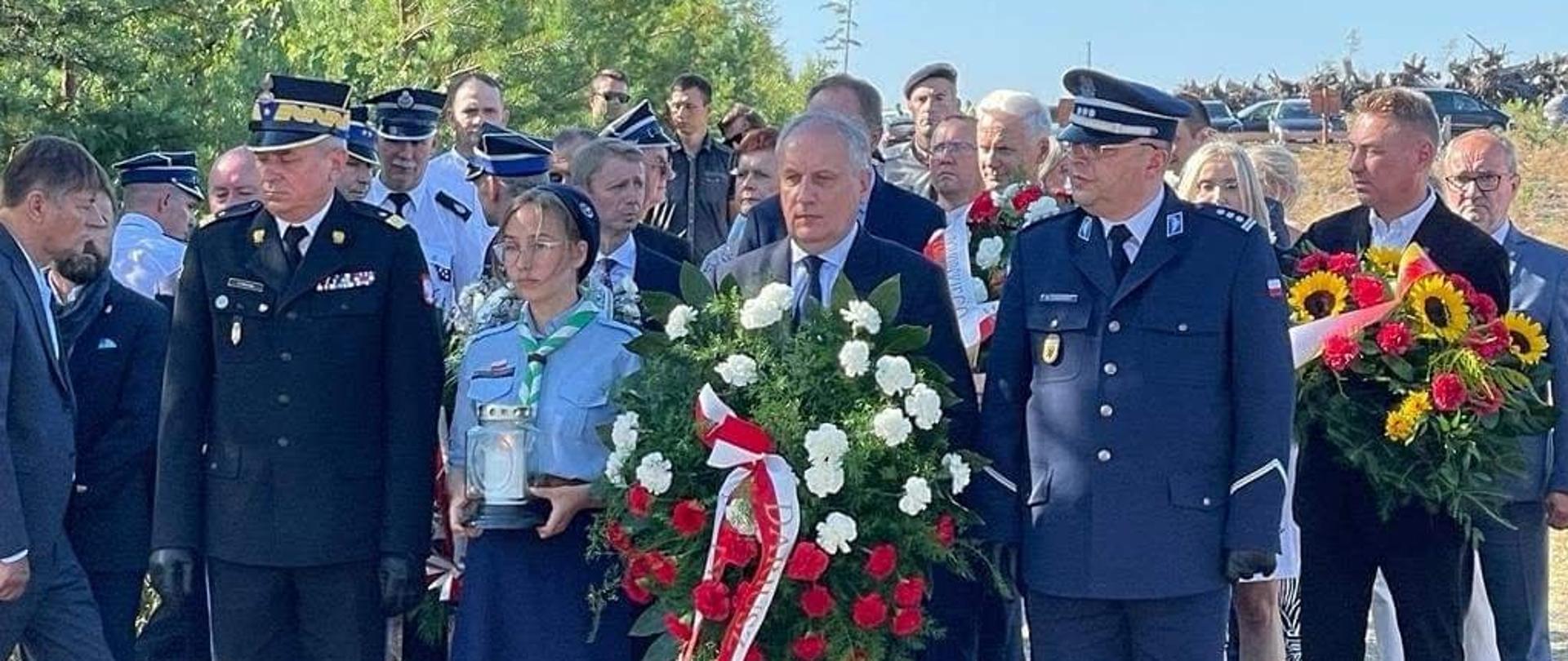 Wojewoda pomorski, harcerka, przedstawiciele służb mundurowych oraz zaproszeni goście trzymają kwiaty stojąc w rzędach obok siebie. 