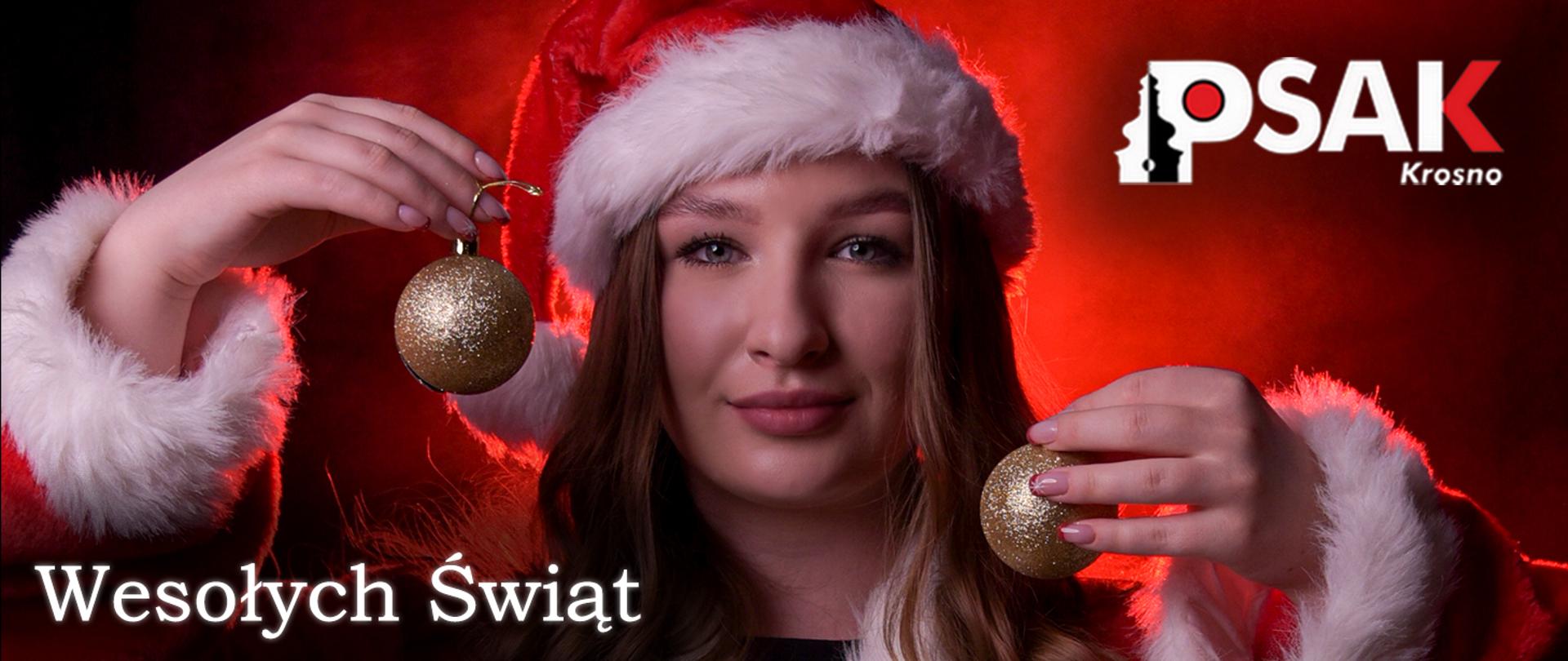  Zdjęcie dziewczyny ubranej w strój Mikołaja na czerwonym tle. Na głowie ma czapkę Mikołaja w dłoniach trzyma złote bombki. 