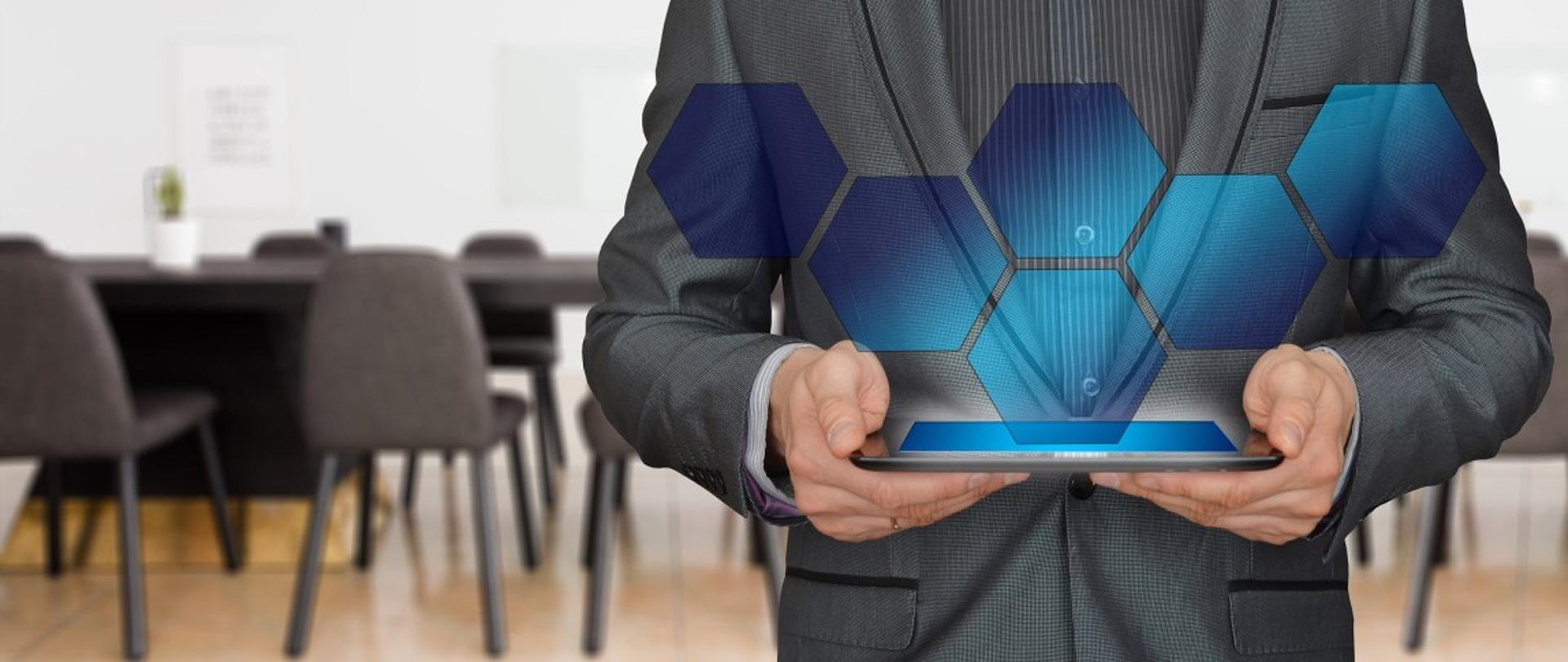 Osoba w garniturze trzymająca tablet z cyfrową grafiką, w tle sala spotkań