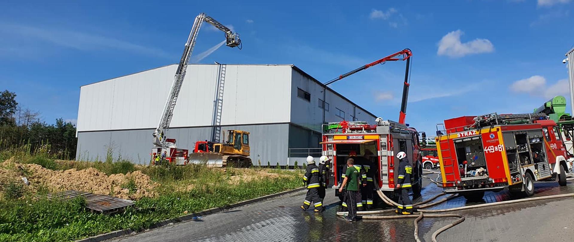 Zdjęcie przedstawia biało – szara halę produkcyjną, przy której rozstawione są 2 strażackie podnośniki. Z ich podestów podawana jest woda na pokrycie dachowe. Przed halą stoją czerwone, ciężarowe samochody pożarnicze z podłączonymi wężami. Koło nich pracują strażacy w ubraniach specjalnych i hełmach.