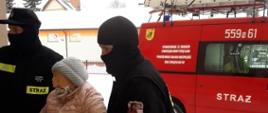 Zdjęcie przedstawia ratowników z Ochotniczej Straży Pożarnej w Spychowie, którzy pomagają starszej kobiecie dostać się po szczepieniu do miejsca zamieszkania.