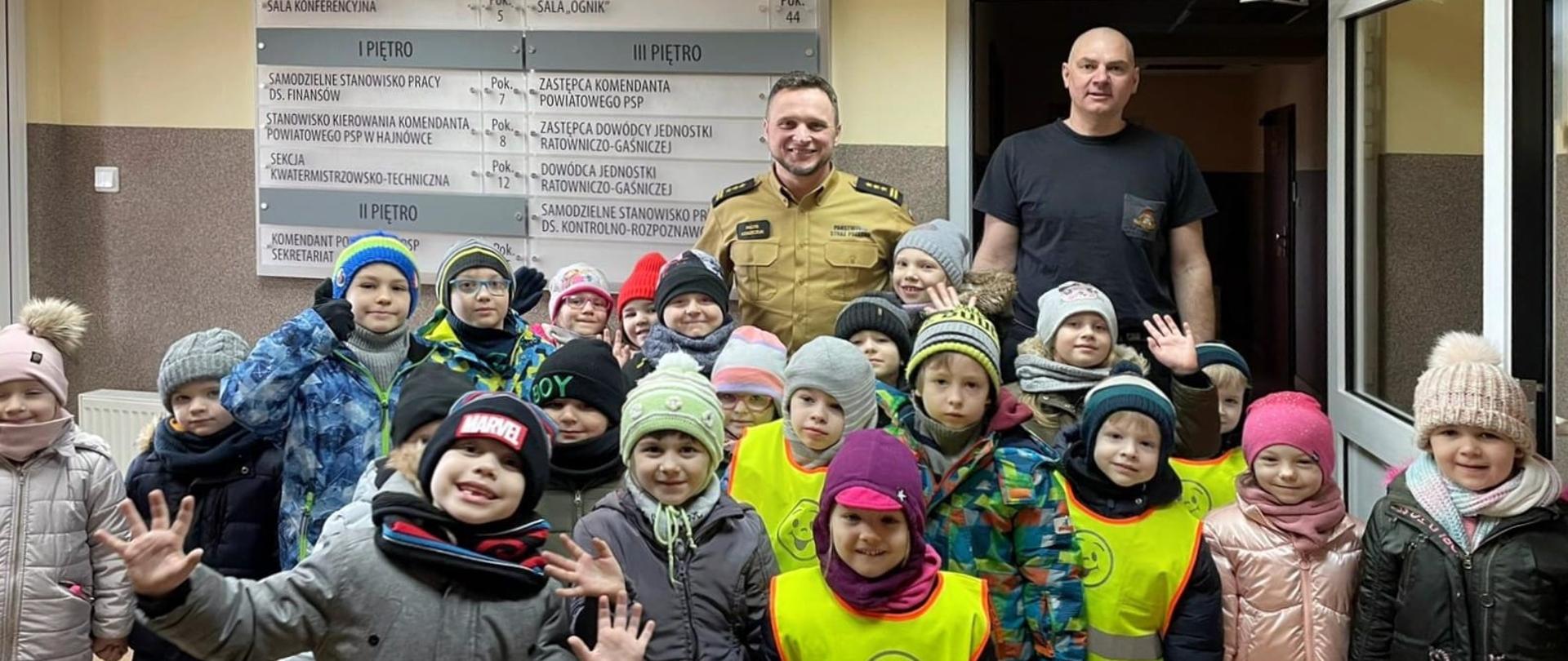 W holu stoi kilkadziesiąt ubranych dzieci. Za nimi dwóch strażaków.