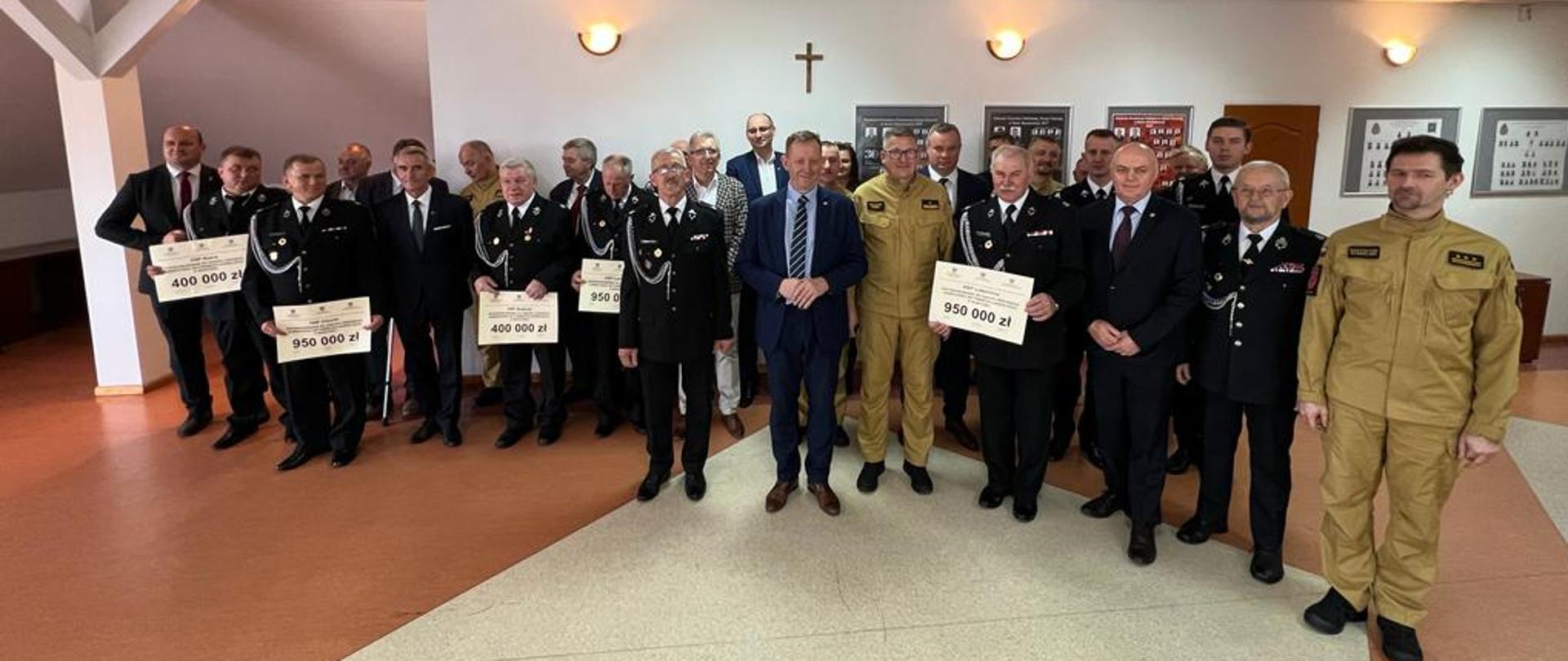 Na zdjęciu druhowie OSP oraz strażacy PSP ubrani w mundury służbowe oraz galowe i pozostali zaproszeni goście stoją ustawieni obok siebie. Pięciu z druhów trzyma w dłoni promesy z wpisaną kwotą promes.