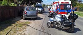 Zdjęcie przedstawia scenerię na miejscu zdarzenia drogowego, do którego doszło na lokalnej drodze w terenie zabudowanym. Po prawej stronie widać dwa zaparkowane motocykle ratunkowe oraz tył karetki pogotowia z włączonymi sygnałami świetlnymi