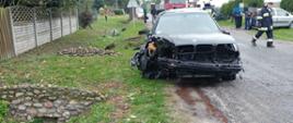 Samochód osobowy marki BMW stoi częściowo na poboczu i na drodze. Ma wgnieciony przód. Za nim leży koło, które odpadło podczas zderzenia. Stoi również za nim wóz strażacki. Z prawej strony strażak idzie ulicą.