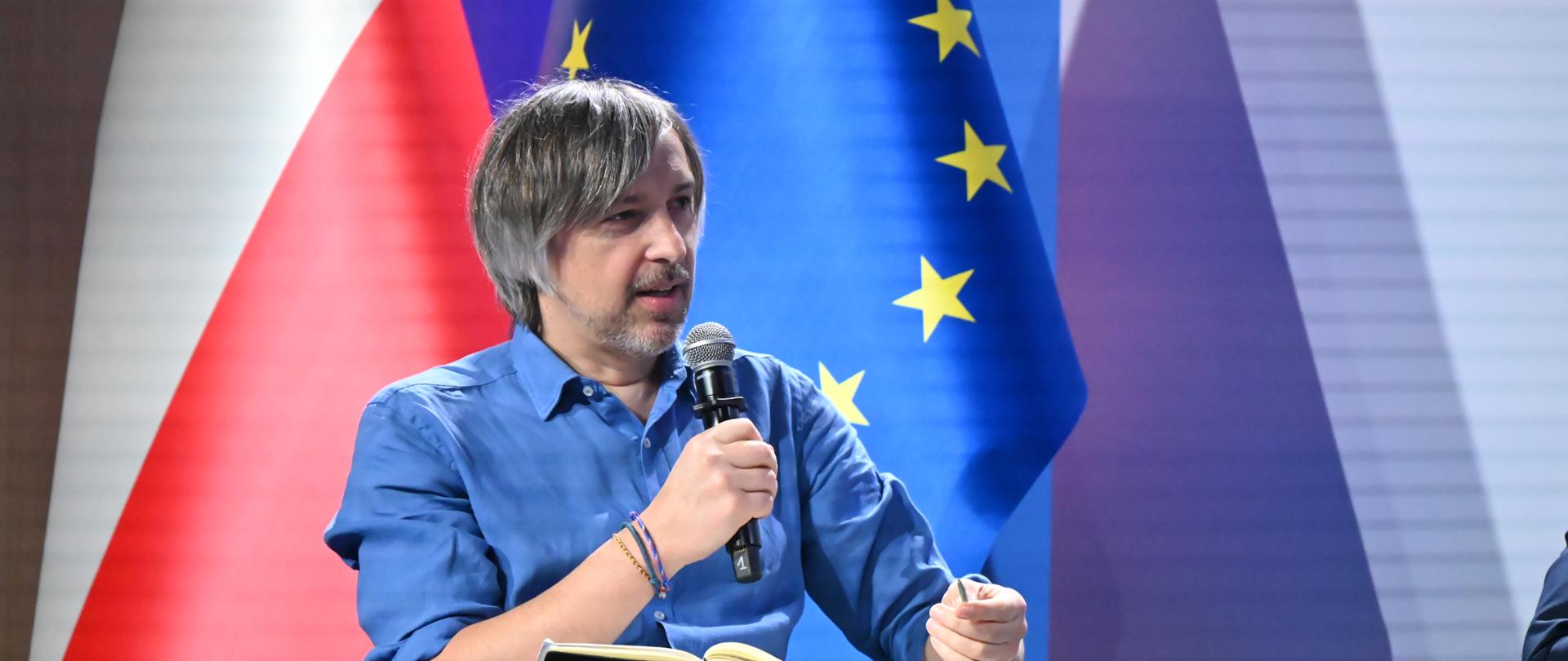 Wiceminister Krzysztof Bolesta podczas konferencji „Rozwój po europejsku. Jaka polityka rozwoju dla bezpiecznej, odpornej i globalnie konkurencyjnej Unii Europejskiej?”. Wiceminister siedzi i mówi do mikrofonu, za nim widać flagę Polski i flagę Unii Europejskiej.