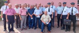 Kolorowe zdjęcie przedstawia grupę kilkunastu osób na zewnątrz przy ścianie budynku. Większość to osoby w ubraniach cywilnych, kilka w mundurach Ochotniczej Straży Pożarnej wraz z Zastępcą Komendanta Powiatowego PSP po wręczeniu decyzji o przyznaniu świadczenia ratowniczego.