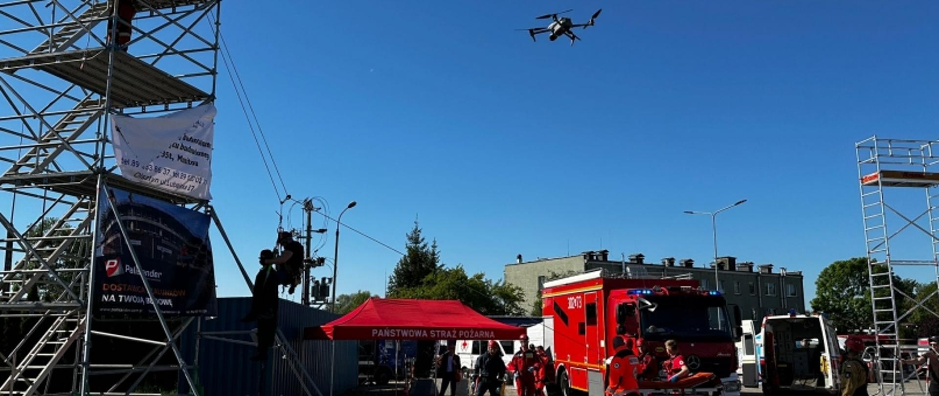 Scena plenerowa z wozem strażackim, dronem, namiotem Państwowej Straży Pożarnej i rusztowaniami; wokół uczestnicy wydarzenia.
