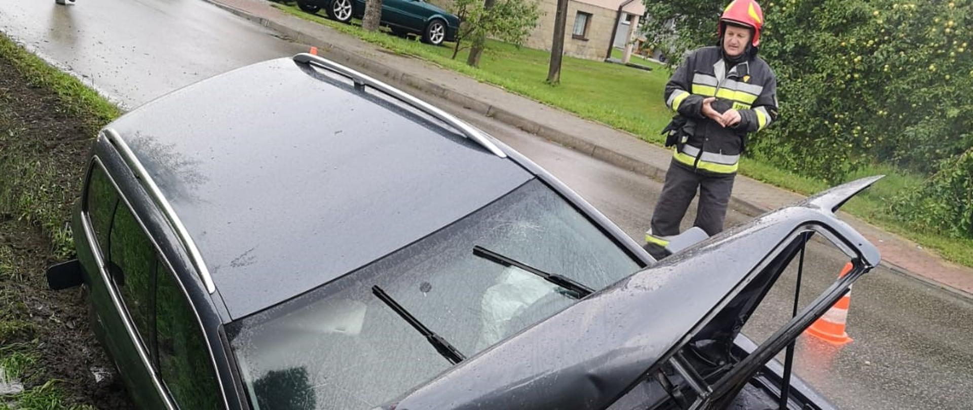 Na zdjęciu widoczny jest samochód osobowy, który w wyniku zjechania do rowu uderzył następnie w betonowy przepust. Powstały zniszczenia przedniej części. Widoczna jezdnia nad pojazdem na której znajdują się strażacy i zabezpieczenie miejsca zdarzenia. W tle zabudowania i roślinność miejscowości.