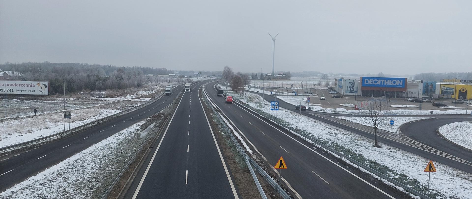 Droga krajowa DK91 zimą, widok z góry na dwupasmową jezdnię prowadzącą z Częstochowy w kierunku Nowej Wsi i dalej na Katowice. Po prawej stronie widoczne sklepy, w tle wiatrak oraz lekko ośnieżone pola.