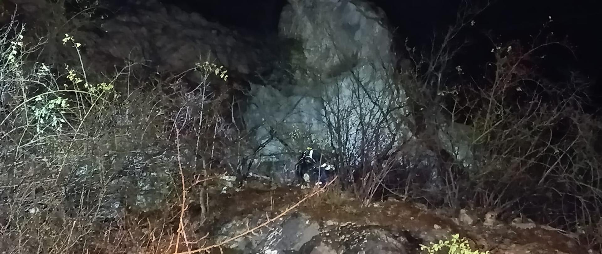 Zdjęcie przedstawia fragment kamieniołomu. Pod skałą, na półce skalnej słabo widoczni pochyleni nad poszkodowaną znajdują się ratownicy. 