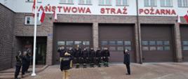 Ujęcia z chwili podnoszenia flagi lub składania honorów. Oficerowie przy maszcie (z lewej) oraz oficer na prawej flance salutują (podnoszą prawą dłoń do nakrycia głowy). Strażacy w szeregu również salutują. Napis na fasadzie jest w pełni widoczny: "PAŃSTWOWA STRAŻ POŻARNA".
