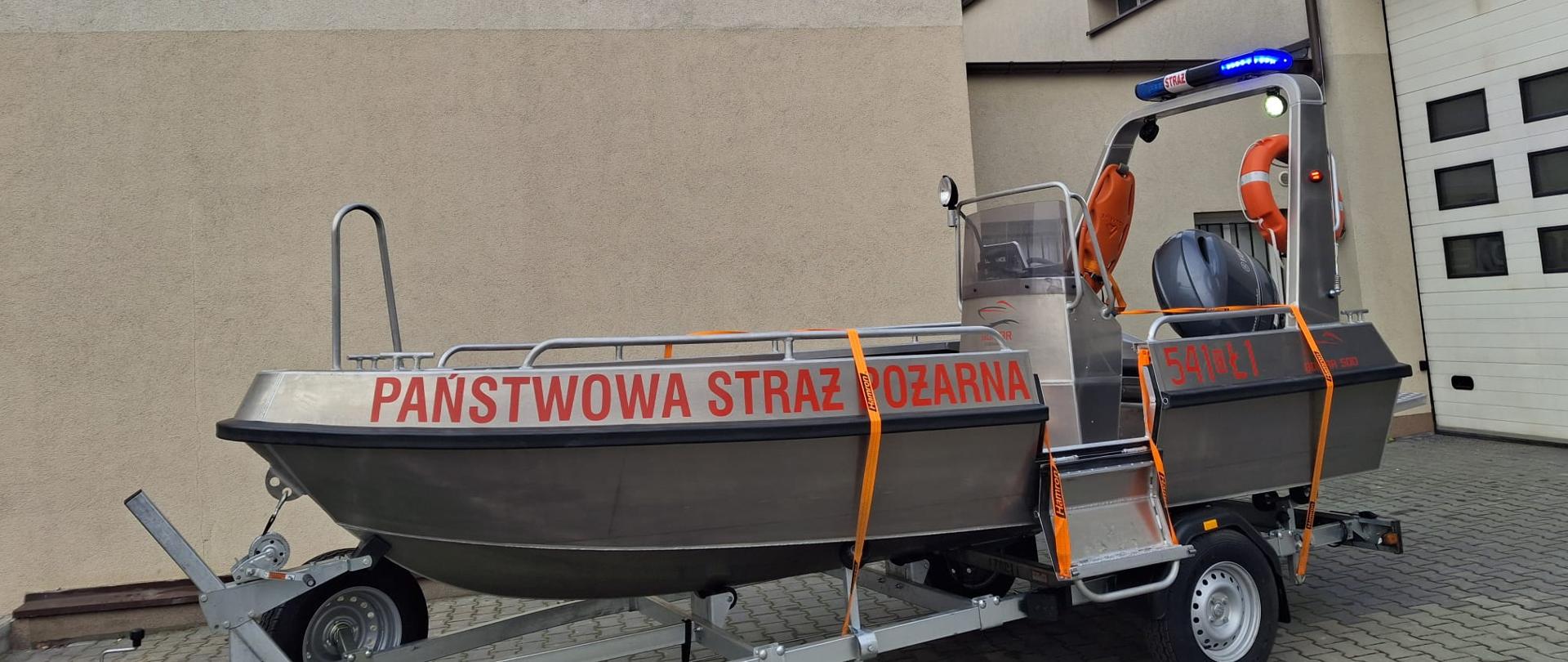 Srebrna łódź ratownicza z napisem PAŃSTWOWA STRAŻ POŻARNA na tle budynku jednostki