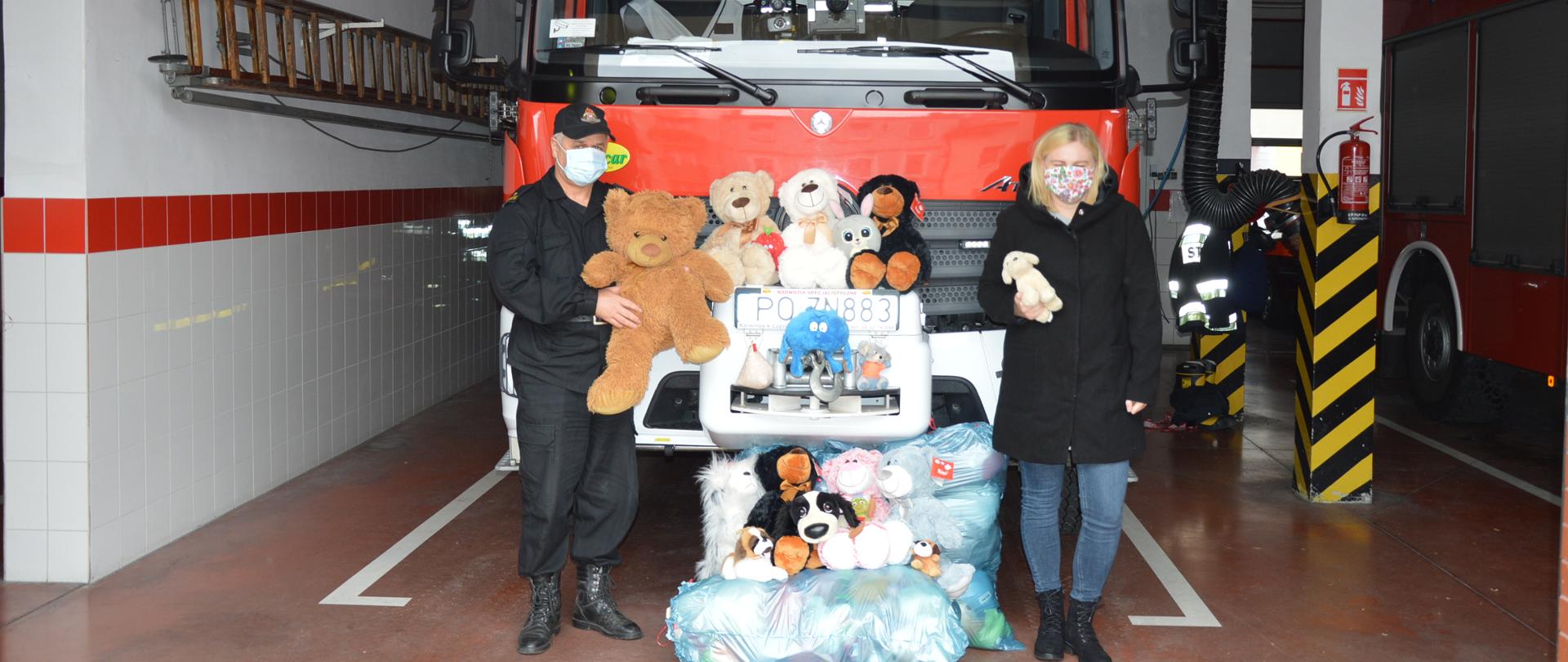 Boks garażowy. W nim wóz strażacki ustawiony przodem do wyjazdu. Na tle pojazdu - z lewej strony strażak trzymający dużą maskotkę misia, z prawej strony - kobieta z mniejszą maskotką w prawej ręce. Pomiędzy nimi ułożone na i przed samochodem wiele innych pluszowych maskotek. Na ścianie z lewej strony samochodu znajduje się zawieszona drabina, na filarze po prawej stronie - gaśnica.