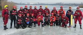 Grupa 16 strażaków PSP z komend powiatowych i miejskiej PSP w Opolu pozuje na lodzie podczas szkolenia doskonalącego z ratownictwa lodowego. Uczestnicy, ubrani w czerwone kombinezony i wyposażeni w sprzęt ratowniczy, doskonalą techniki poruszania się po lodzie oraz ewakuacji poszkodowanych w warunkach kruchego lodu.