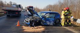 Rozbity niebieski samochód osobowy stojący w poprzek drogi, obok niego dwóch strażaków, na drodze za nim inne auta osobowe i ciężarowe, w prawej części zdjęcia las.