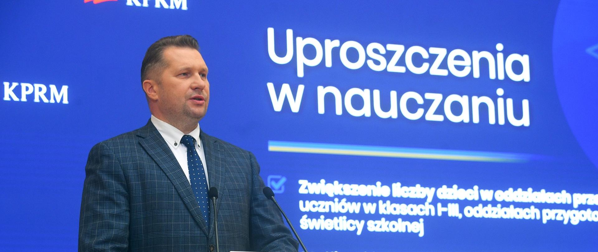 Minister Czarnek przemawia do mikrofonu na tle niebieskiej ściany z napisem Uproszczenia w nauczaniu.