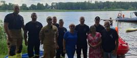Grupa strażaków i mieszkańców pozuje do wspólnego zdjęcia nad brzegiem jeziora, w tle łodzie służb ratunkowych i wypoczywający ludzie.