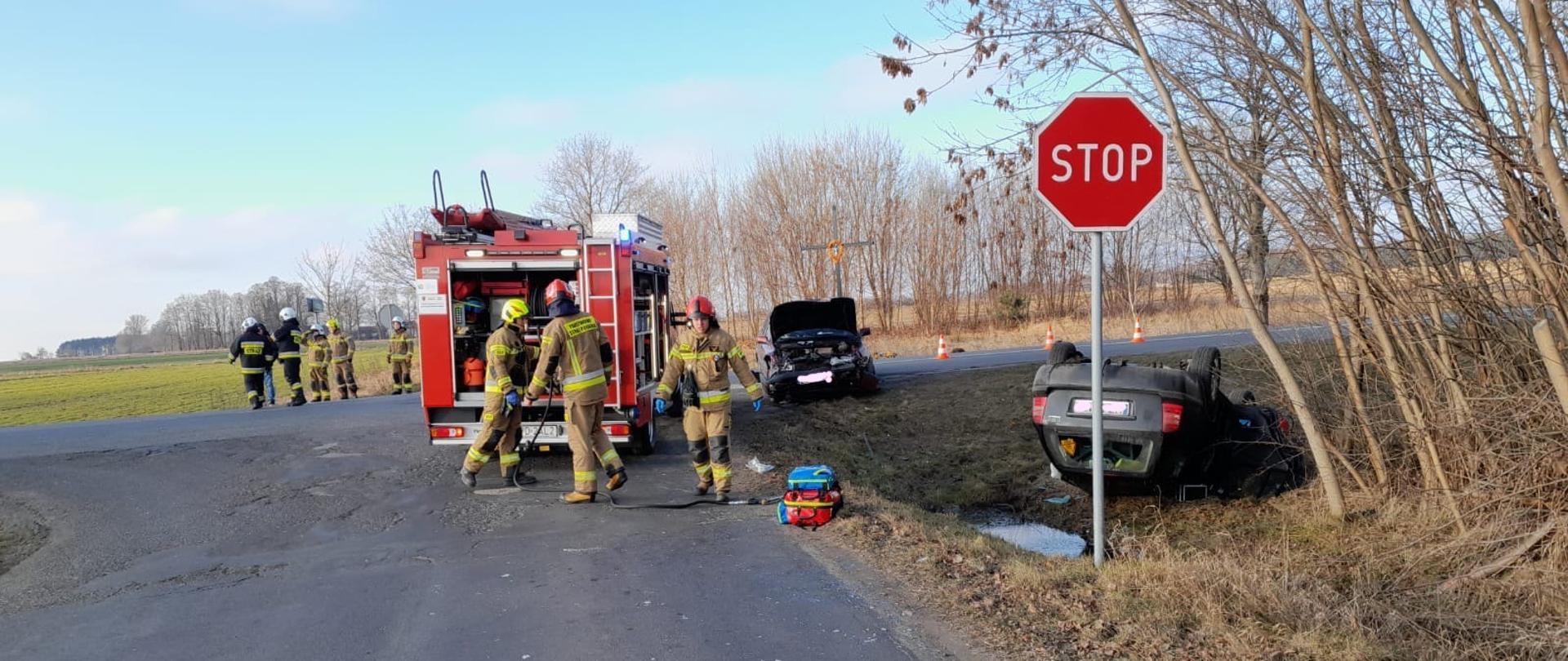 po prawej stronie w rowie leży wywrócony na dach ciemny samochód osobowy z wybitą tylną szybą, na asfalcie stoi wóz strażacki obok niego strażacy z przodu stoi drugie rozbite auto osobowe z podniesioną do góry maską