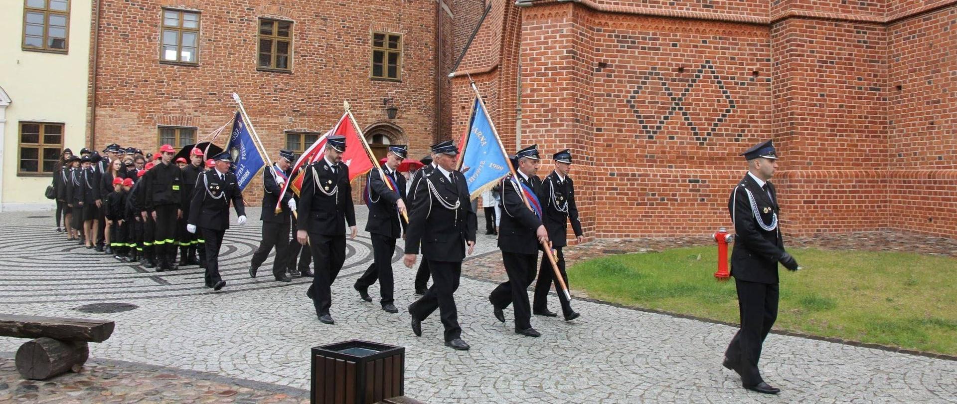 Strażacy w mundurach galowych idą chodnikiem. Na czele trzy sztandary. W tle kościół z czerwonej cegły.