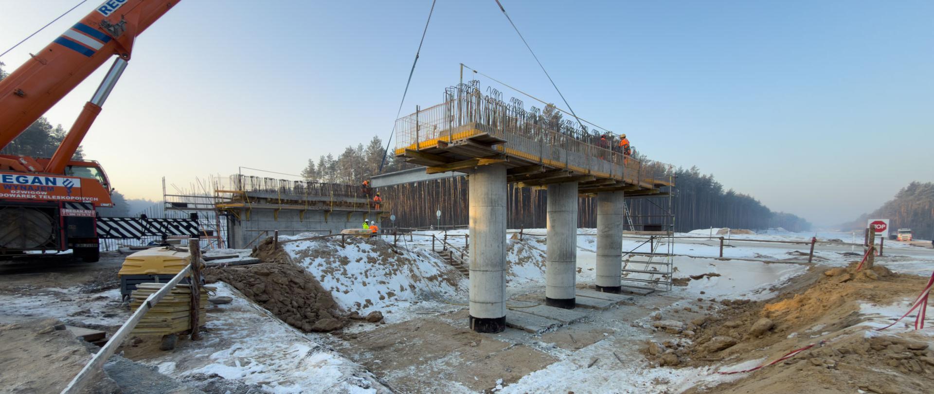 Budowa obiektu mostowego z trzema betonowymi podporami oraz konstrukcją pomostu podnoszoną przez dźwig.