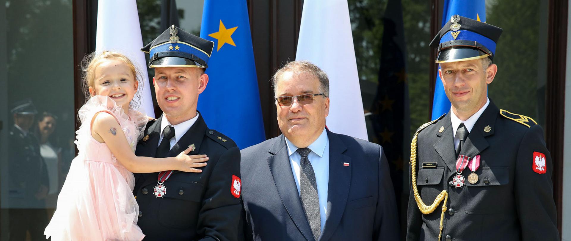 Po lewej stronie strażak w mundurze wyjściowym z dziewczynką w jasnej sukience na rękach. Na środku Minister Andrzej Dera w garniturze. Po prawej strażak w mundurze wyjściowym. Na mundurach obu strażaków widoczne Krzyże zasługi za dzielność. W tle flagi Polski i Unii Europejskiej