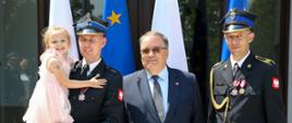 Po lewej stronie strażak w mundurze wyjściowym z dziewczynką w jasnej sukience na rękach. Na środku Minister Andrzej Dera w garniturze. Po prawej strażak w mundurze wyjściowym. Na mundurach obu strażaków widoczne Krzyże zasługi za dzielność. W tle flagi Polski i Unii Europejskiej
