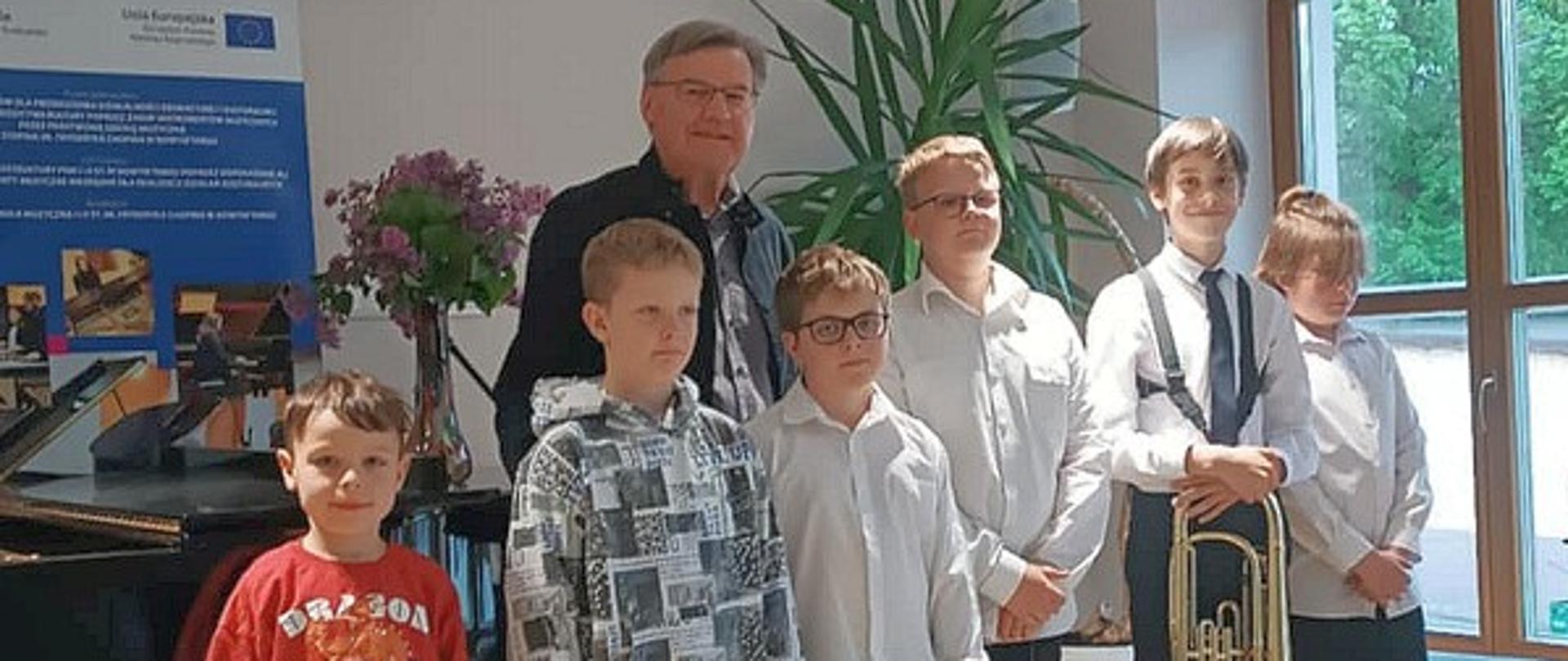 Fotografia przedstawiająca uczniów Państwowej Szkoły Muzycznej I i II stopnia im. F.Chopina w Nowym Targu filia w Jaworkach ze swoim nauczycielem P Józefem Pańszczykiem. Na pierwszym planie stojący na podwyższeniu sceny 6 chłopców, jeden z nich trzymający swój instrument. Za nimi nauczyciel. W tle widoczny czarny fortepian ze stojącym w szklanym wazonie bukiet fioletowych kwiatów bzu oraz zielony wysoki kwiat juki.