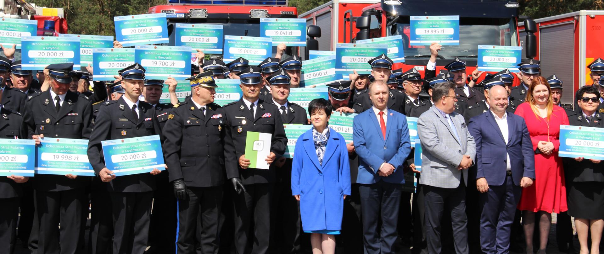 Grupowe zdjęcie druhów, parlamentarzystów i symbolicznych voucherów trzymanych w dłoniach przez kilkudziesięciu druhów. W tle pojazdy pożarnicze.