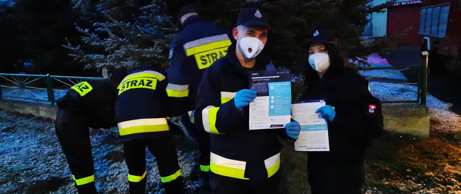 Strażacy ochotnicy prezentujący roznoszone materiały informacyjne na temat profilaktyki oraz szczepień przeciwko COVID-19 na tle zabudowań wiejskich